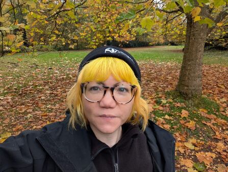 A woman with short dyed yellow hair is looking at the camera. She is wearing tortoishell glasses and a black beanie. She is stood in a park during autumn, surrounded by fallen leaves.
