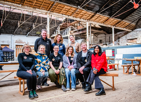 6 of the staff team from the environmental leadership programme are sat on a bench in the middle of a warehouse, with three members stood behind. They are dressed in casual clothing and smiling at the camera.