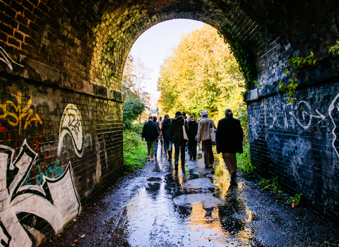 A group of young people are walking away from the camera, out of a brick graffitied tunnel. Out of the tunnel's exit are green trees and bushes in bright sunlight.
