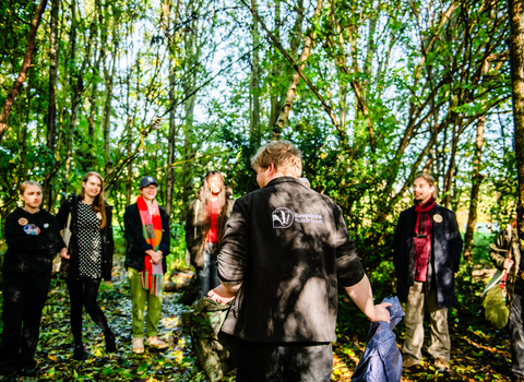 A group of young people are gathered in front of a staff member from Shropshire Wildlife Trusts, who is stood with his back to the camera, the logo of the trust visible on the back of his fleece. There are five young people facing the camera, but out of focus, with their attention focused on the staff member. They are stood in a wooded area, with tall trees with green leaves in bright sunlight.