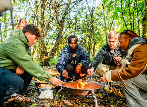 Four young people are crouched around a campfire, toasting marshmallows on sticks. They are surrounded by trees with green leaves on a sunny day. 